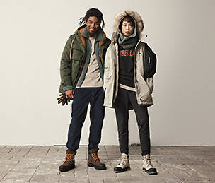 Image of a young woman and young man looking directly at the camera, standing on top of stone pavers. The man has his right arm over the shoulder of the woman and both are in a relaxed pose wearing Timberland winter gear including Timberland jackets, Timberland sweatpants and Timberland boots.