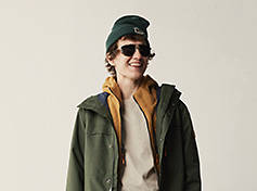 Image of a young man with a green winter hat, green Timberland 3-in-1 winter coat and sunglasses. He is facing down with his right hand on the back of his head.