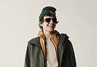 Image of a young man with a green winter hat, green Timberland 3-in-1 winter coat and sunglasses. He is facing down with his right hand on the back of his head.