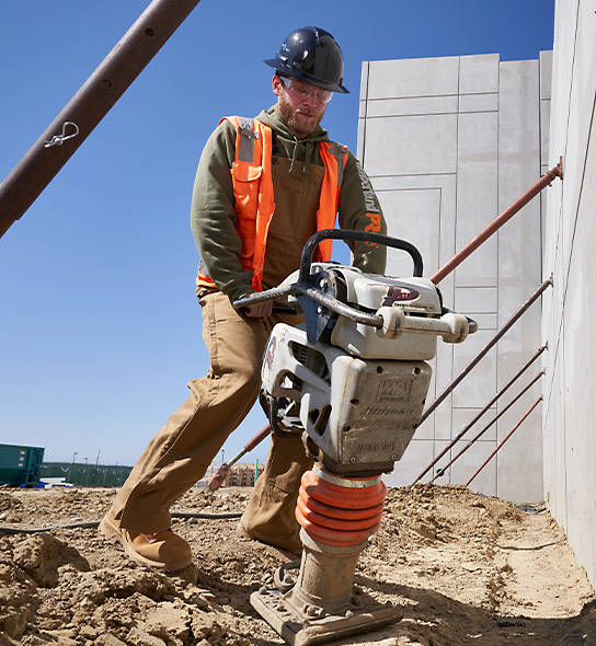 Image of a male construction worker using a large tool to compact dirt while wearing Timberland PRO clothing along with Timberland PRO boots on a clear, sunny day.