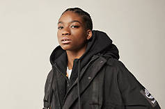 Image of the left-side of a woman in braids, cropped from the chest and above, against a light grey background, wearing a black Timberland jacket.