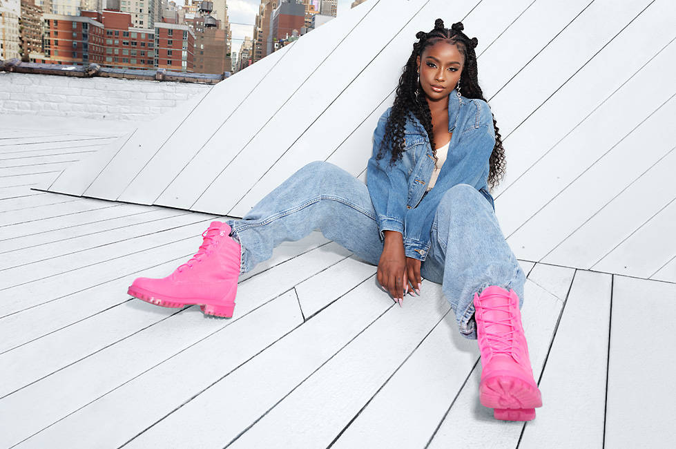 Image of a woman leaning against an all white painted, white roof section at the top of an apartment building in New York, with several brick apartment building in the background. She is wearing a white tank top with a lightweight denim coat along with denim baggy jeans. To top off the outfit, her feat are outfitted with bright pink Timberland X Jimmy Choo boots.