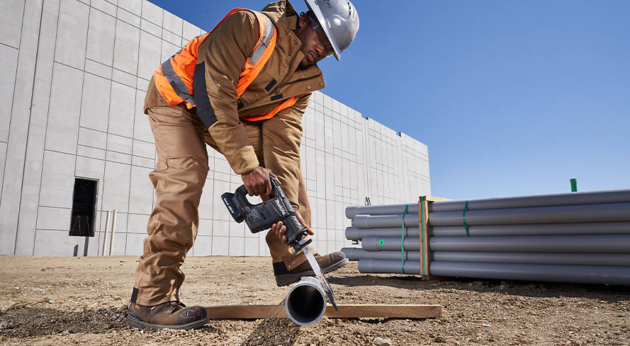 Image of a male construction worker cutting through PVC pipe with an electric saw. He is outside of the warehouse his team is constructing on a clear, sunny day while wearing all Timberland PRO outerwear and Timberland PRO boots.