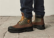 Image of a pair of MEN'S TIMBERLAND® PREMIUM 6-INCH WATERPROOF RUBBER-TOE BOOTS with a color of medium brown nubuck, pictured with a pair of denim jeans cropped from the shins down. The model is standing on light gray paver stones.