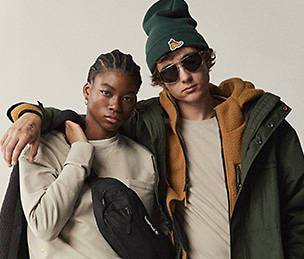 Image of a young adult female with braids on the left and a young male with brown hair on the right, both of which are looking straight into the camera pictured from the waist and above. The female is holding a black jacket over her right shoulder while wearing an light beige crewneck sweatshirt and a black crossbody bag. The male is wearing a dark green winter skully hat and a Timberland 3-in-1 jacket with a light beige t-shirt underneath. All clothing is Timberland brand.