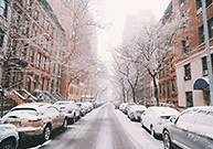 Image of a city street lined with brownstone buildings and parked cars, recently dusted with snow.