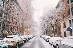 Image of a city street lined with brownstone buildings and parked cars, recently dusted with snow.