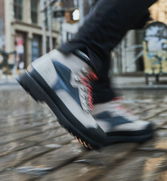 Motion shot of an individual walking with these boots on a city's cobblestone streets showing a cropped image from the shin down to the sidewalk