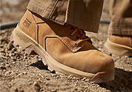 Close up image of the left foot of a worker on a pile of dirt. The worker is wearing Timberland Men's Direct Attach 6-inch Steel Toe Waterproof Work Boots in the color of wheat nubuck.