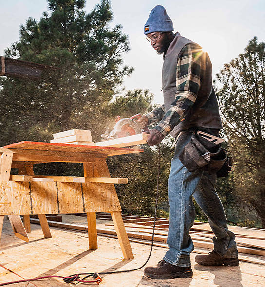 A male construction worker is using a circular saw to cut a 2x4 in the outdoors. It appears to be close to sunset and is sunny outside with pine trees in the background. The worker is standing upright while wearing almost all Timberland PRO clothing including a light blue Timberland PRO® Essential Watch Cap, a Men's Timberland PRO® Woodfort Midweight Flannel Work Shirt which has a dark green and light yellow pattern, blue jeans and Timberland PRO workboots.