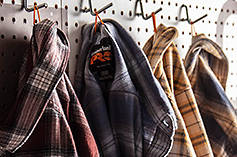 Image of four different Men's Timberland PRO® Woodfort Midweight Flannel Work Shirts hanging on metal hooks in a workshop.