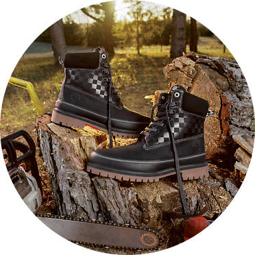 Image of a pair of Vans X Timberland black boots with the Vans checkered pattern on the upper half and brown soles. Boots are sitting on top of wooden tree stumps with the sun setting in the background and a chainsaw in the foreground.