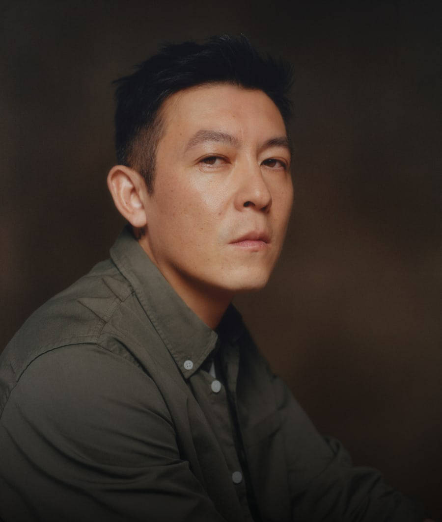 Edison Chen regardant la caméra, de la poitrine vers le haut dans une chemise verte boutonnée sur un fond brun.