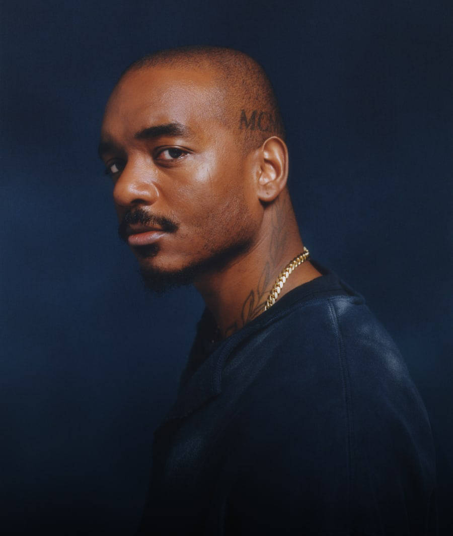 Samuel Ross looking left at the camera, from chest up in a blue sweatshirt with a gold chain necklace against a blue background.