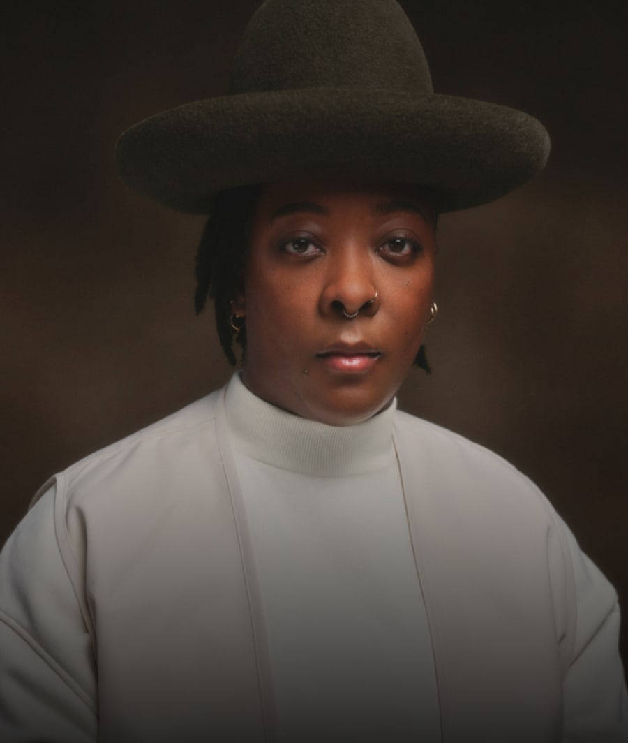 Nina Chanel Abney looking straight ahead at the camera, from chest up in a white shirt with a light tan vest and brown hat against a brown background.