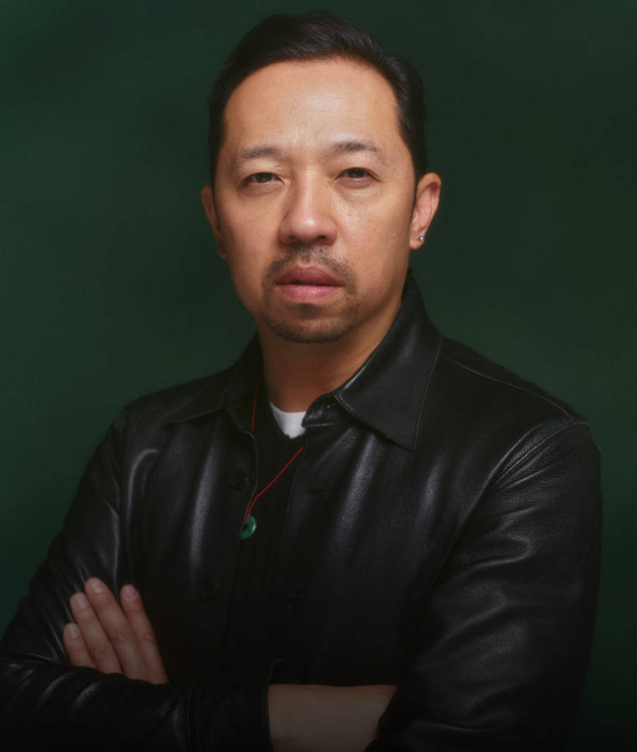 Humberto Leon looking straight ahead at the camera with his arms crossed over his chest, from chest up in a black leather jacket over a black v-neck and white t-shirt with a green circular pendant on a red cord around his neck against a hunter green background.