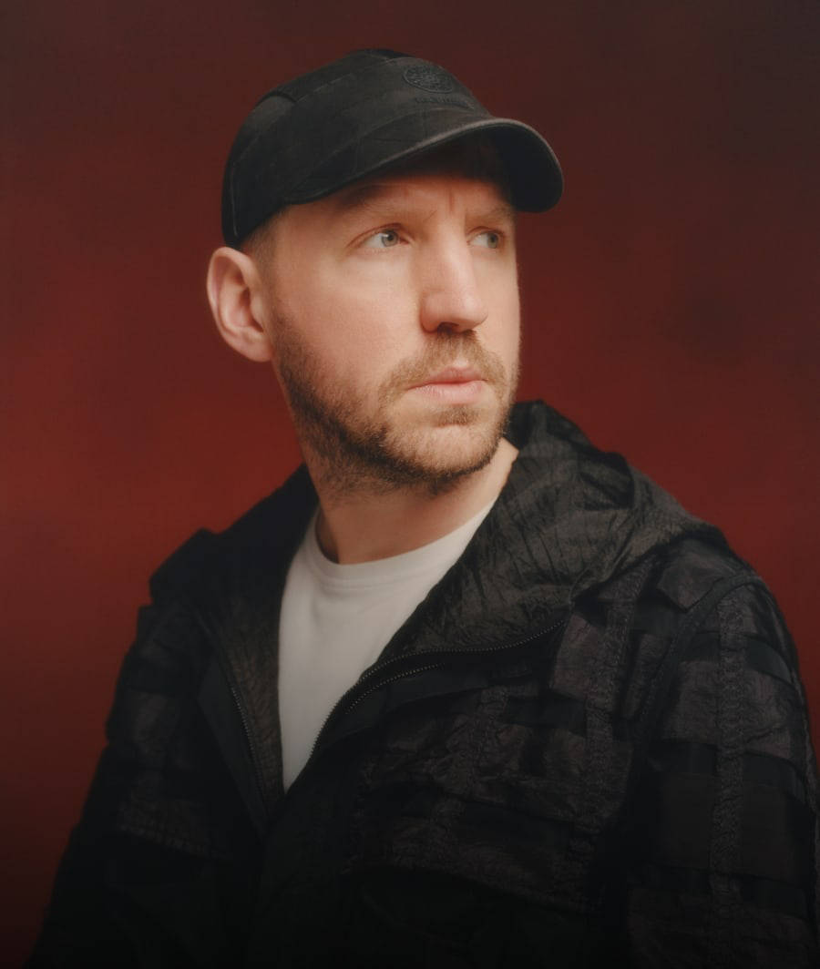Christopher Raeburn looking to the viewer's right, from the chest up in a black jacket over a white t-shirt with a black hat against an orange-red background.