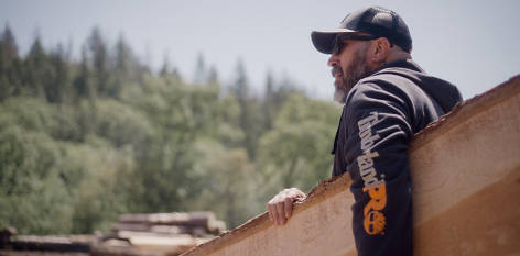 Image of Josh in a gray Timberland PRO baseball cap, sunglasses and navy blue Timberland PRO hooded sweatshirt, carrying a large slab of wood.
