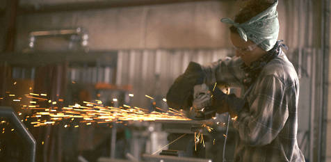 Image of Kelli wearing safety goggles, a Timberland PRO gray and blue flannel shirt as she welds while bright sparks shoot up into the air.