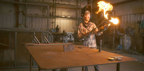 Image of Kelli at a welding table, wearing safety goggles, a Timberland PRO gray and blue flannel shirt as she lifts a fiery welding torch into the air.