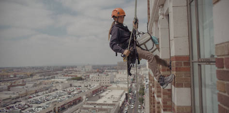 Image of Natalia in an orange hard hat and blue Timberland PRO sweatshirt in profile,  showing her entire body in action as she rappels down the side of a tall building.