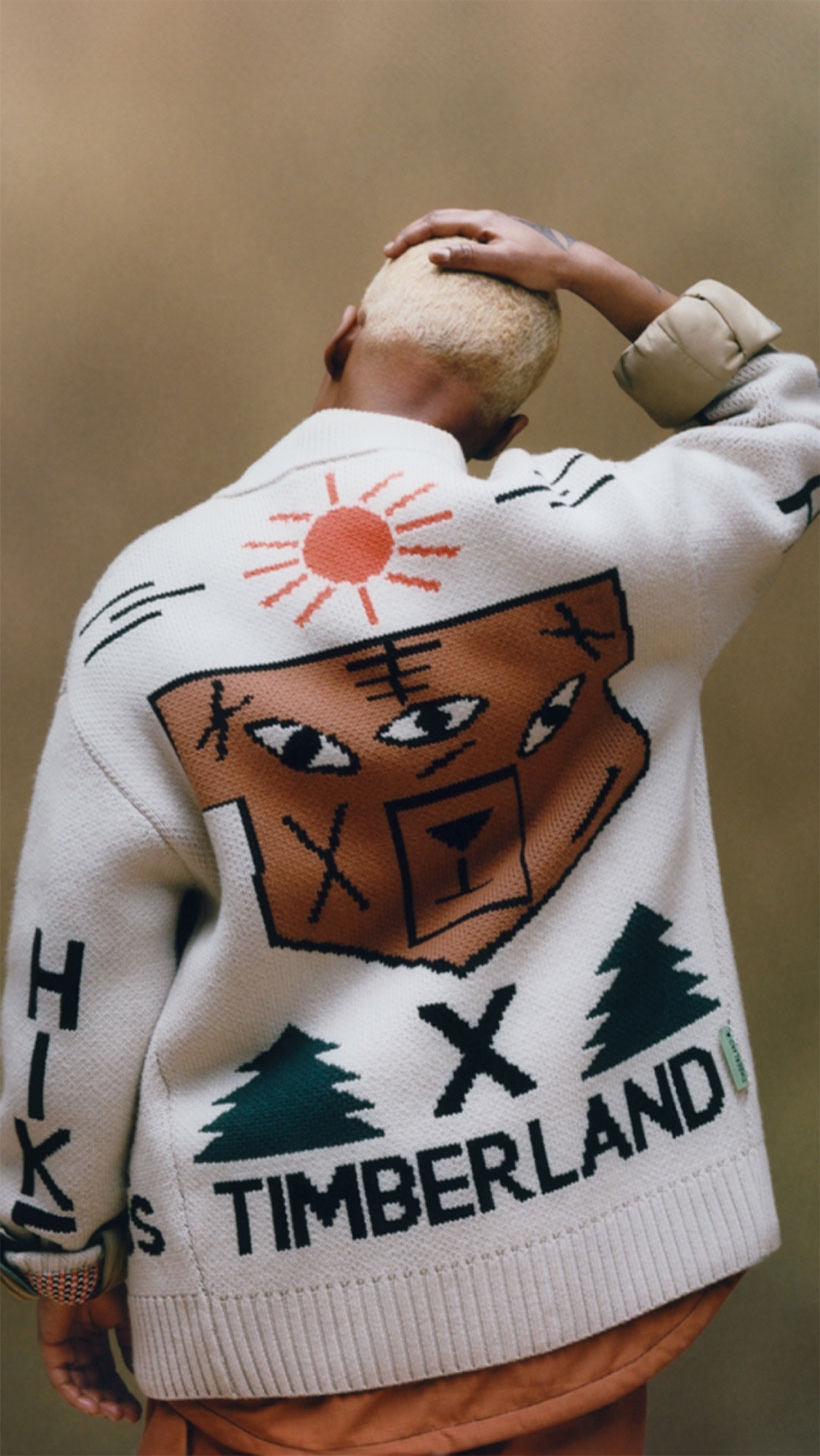 A standing person with their right hand on their head, photographed from the waist up from behind, wearing a white cardigan with a knit design of an orange sun, a brown shape containing three eye-like shapes and several black shapes above a black X between green pine tree shapes, above the word TIMBERLAND in black. The word HIKE is knitted into the arms.