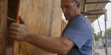 Image of Roger in a blue Timberland PRO t-shirt, inside a new construction framed structure, looking at some plans with a tape measure.