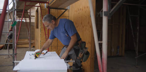 Image of Roger in a blue Timberland PRO t-shirt, inside a new construction framed structure, wearing safety goggles and carrying a large board of plywood.